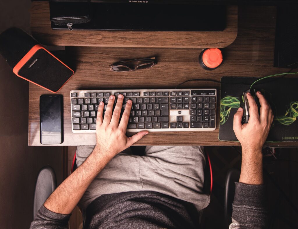 A top-down view of a man using a desktop computer with various electronics on a wooden desk.